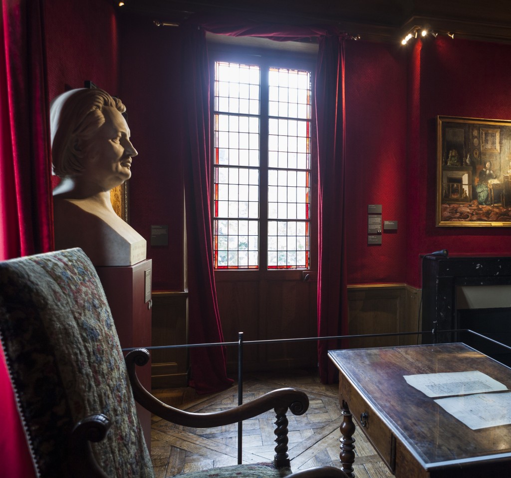 Dark interior view of Balzac's office: dark wood desk with manuscript on it, in front of a bust of Balzac and with dark red wall and window in the background
