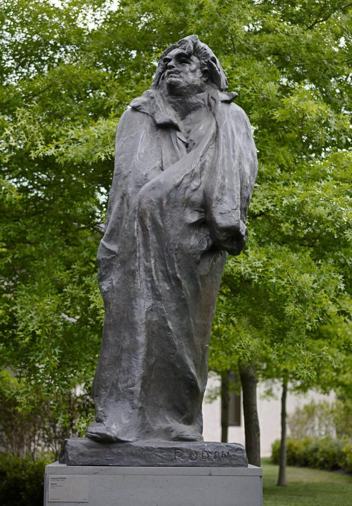 Large, darkened bronze sculpture of imposing male figure wrapped in a dramatic cloak, against a leafy tree background