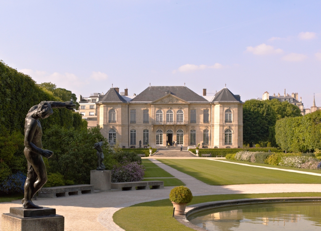 Sunny view of a stately French mansion, with a long lawn, round pond and nude bronze sculpture reaching forward